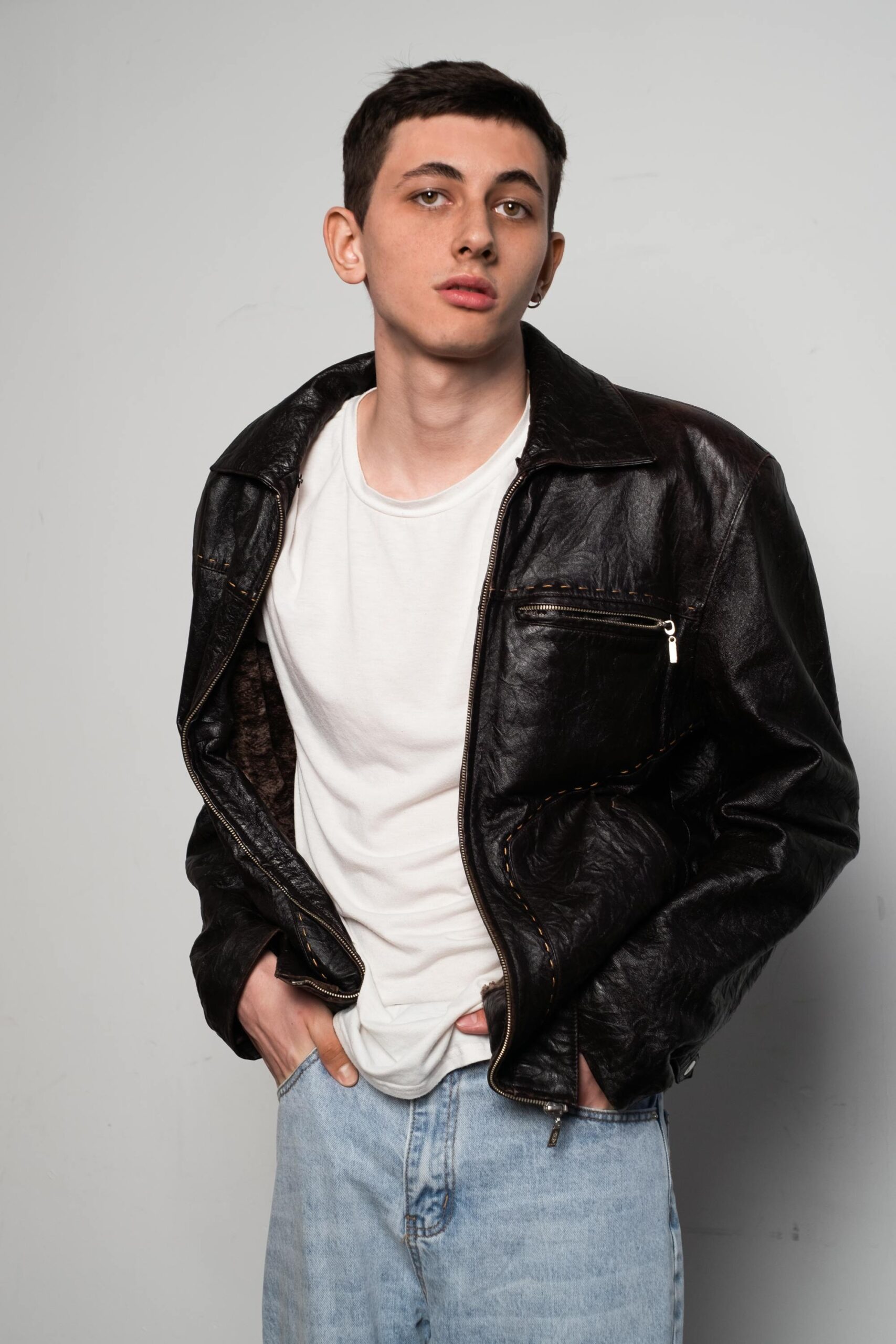 Stylish studio portrait of a man wearing a black leather jacket and white shirt, hands in pockets.